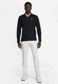 Črna polo majica z dolgimi rokavi in četrtim zadrgo ter belim ovratnikom, v kombinaciji z belimi slim-fit hlačami in belimi čevlji, ki imajo Nike logo.