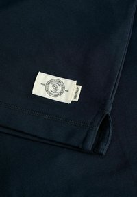 Tissu bleu foncé avec une étiquette de vêtement rectangulaire blanche cousue près d’une fente située sur la couture latérale, affichant les détails de la marque et le texte « Originals ».