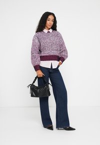 Mulher vestindo um suéter de malha roxo sobre uma camisa rosa, jeans de perna larga, saltos pretos, segurando uma mala de couro preta com franzido.