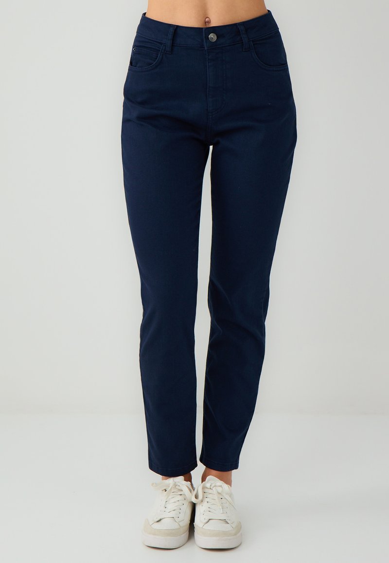 Donna che indossa pantaloni aderenti blu scuro e sneakers bianche, in piedi contro uno sfondo chiaro e semplice.
