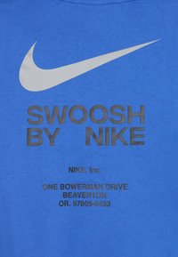 Modra majica iz bombažne mešanice z velikim sivo-črnim logotipom Nike Swoosh in črnim besedilom "SWOOSH BY NIKE" ter naslovom podjetja.