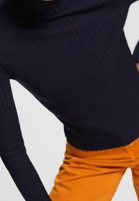 Marineblaues, geripptes Langarmoberteil kombiniert mit leuchtend orangefarbenen Hosen. Das Oberteil verfügt über einen hohen Ausschnitt und schafft eine taillierte Silhouette.