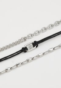 Trois chaînes : une fine chaîne en argent, une chaîne en maillons d'argent plus épaisse, et un cordon tressé noir avec un fermoir métallique. Le tout sur un fond blanc.