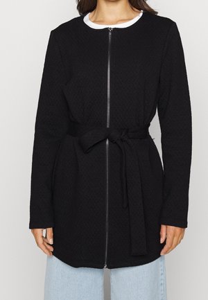 Kobieta w długim rękawie teksturowanej czarnej kurtce z przednim zamkiem błyskawicznym i związanym paskiem z tkaniny, w połączeniu z jasnoniebieskimi dżinsami.