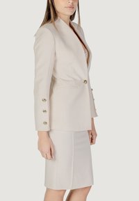 Giacca di pantalone beige chiaro con un design aderente, dettagli in bottone dorato e una gonna a matita coordinata, lunga fino al ginocchio. Realizzata con spalle strutturate.