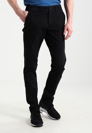 Man die een zwarte slim-fit broek draagt, met zijn hand in zijn zak en zwarte sneakers, staand tegen een eenvoudige lichte achtergrond.