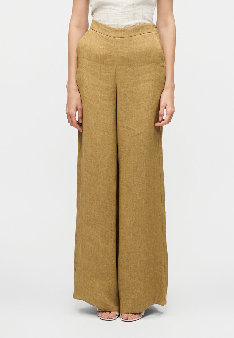 Γυναίκα που φοράει ψηλόμεσο, φαρδύ παντελόνι μουσταρδί από λινό και λευκό αμάνικο τοπ, στέκεται με τα χέρια χαλαρά δίπλα στο σώμα.