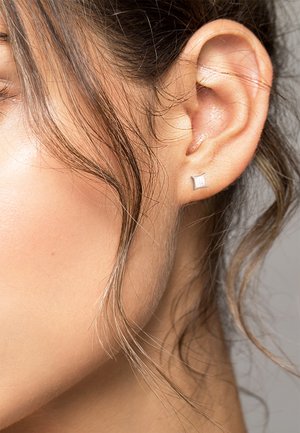 Silberner würfelförmiger Ohrstecker mit strukturiertem Oberflächenfinish. Nahaufnahme, die den Ohrstecker am Ohr zeigt, umrahmt von welligen Haaren, die das Gesicht einrahmen.