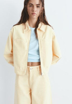 Mladi oseba z dolgimi lasmi nosi bledo rumen jopič in hlače čez svetlo modro kratko majico, stoječa na belem ozadju.