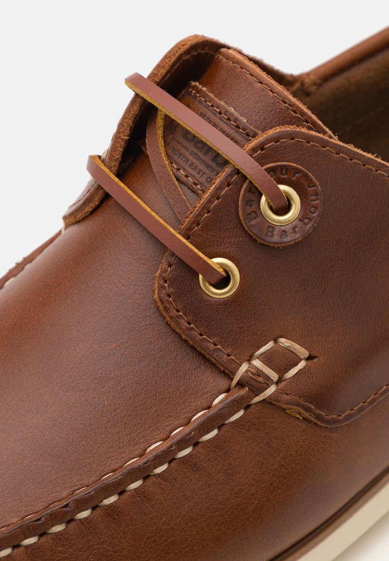 Chaussure bateau en cuir marron avec œillets dorés, lacets contrastants et détails de couture visibles. Elle présente une finition texturée et un emblème de la marque en relief.