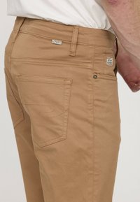 Chinos tan avec une silhouette ajustée, des poches arrière avec des détails de couture, et un accent en métal près de la taille. Tissu lisse et durable.