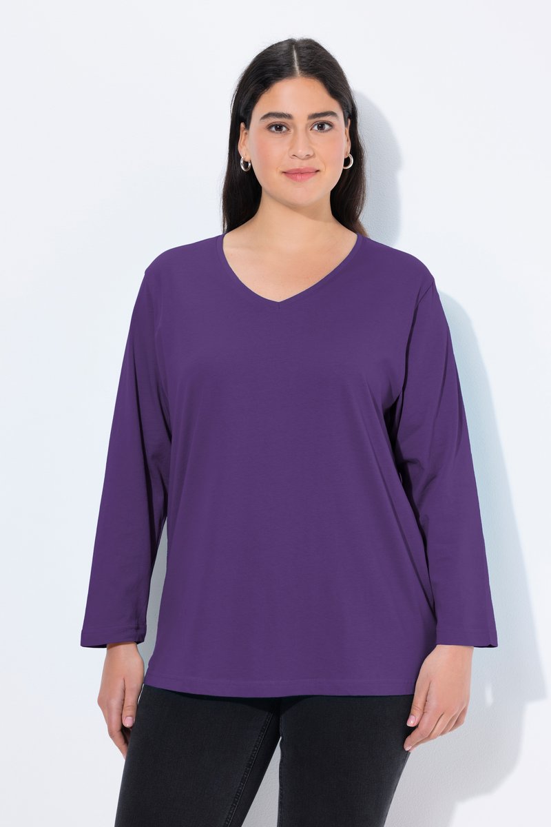 Femme portant un haut violet à manches longues avec col en V et un pantalon noir, debout sur un fond blanc.