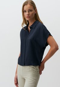 Camicia navy a maniche corte con colletto, realizzata in un tessuto testurizzato. Presenta una chiusura con bottoni e una vestibilità comoda. Abbinata a pantaloni leggeri.