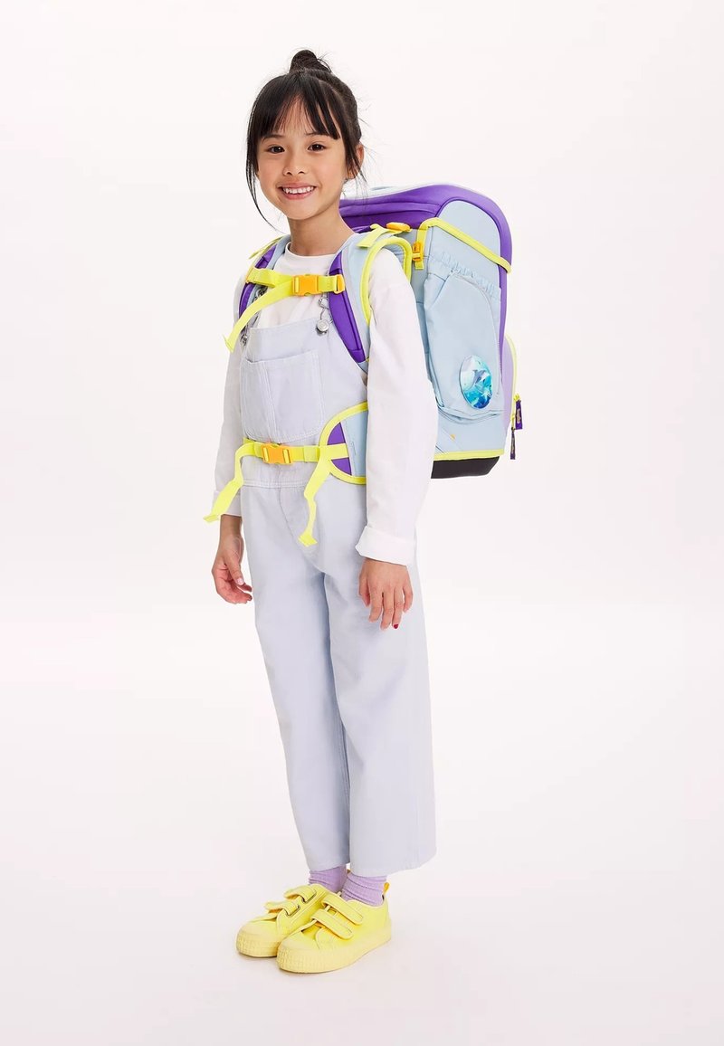 Zaino azzurro chiaro con dettagli viola e cinghie gialle. Il bambino indossa una salopette azzurro chiaro, scarpe gialle e calzini viola.