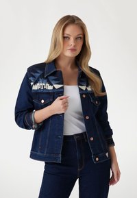 Tamsiai džinsinis švarkas su geometriniu audinių raštu ant pečių, su vario furnitūra, sagomis ir šiek tiek prigludusia forma.