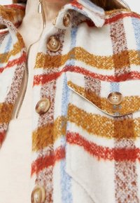 Chaqueta de mezcla de lana a cuadros en blanco, rojo, marrón y azul. Presenta botones grandes y un bolsillo en el pecho con bordes redondeados.
