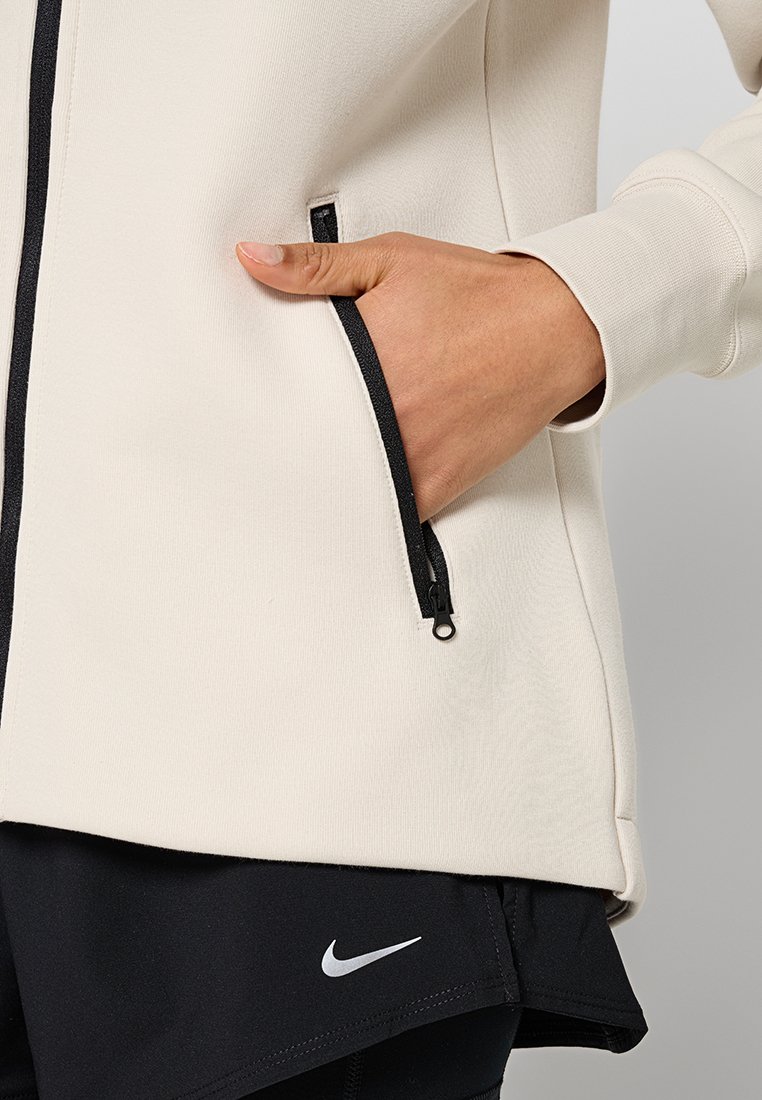 Giacca atletica beige chiaro con tasca con zip, dettagli neri sulla zip e texture liscia; indossata sopra pantaloni corti neri con logo bianco.
