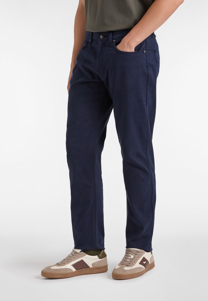 Chinos azul marino con un corte ajustado, que cuentan con un cierre de botón y bolsillos laterales. Combinados con zapatillas beige y oliva, destacando un aspecto texturizado.