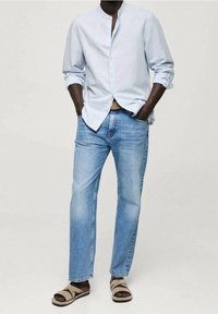 Camicia azzurra chiara con colletto alla mandarina, jeans blu e sandali marroni con cinturini, in piedi con le mani nelle tasche.