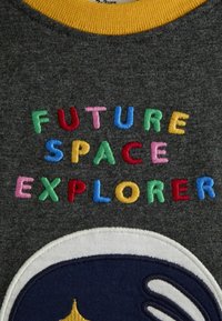 Šedá mikina s žltým golierom; obsahuje viacfarebný vyšívaný text "BUDÚCI PRIEKOPNÍK SPACU" a kruhový tmavomodrý dizajn so žltými akcentmi.