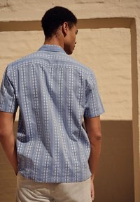 Chemise à manches courtes bleue à motifs avec rayures verticales et accents floraux. Tissu léger à texture lisse. Col classique avec détails de boutons.