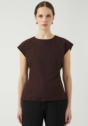 Kobieta ubrana w dopasowany, ciemnobrązowy top bez rękawów i czarne spodnie, stojąca na tle jednolitego jasnego tła, zwrócona przodem.