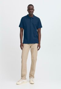 Mann som står iført en marineblå polo, beige bukser og hvite joggesko mot en enkel lys bakgrunn.