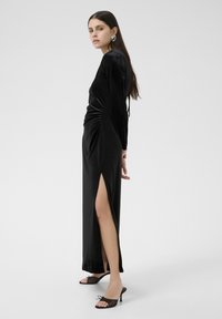 Robe longue en velours noir avec de longues manches, une fente sur le côté et des détails froncés. Portée avec des sandales à talons transparentes et une créole imposante.