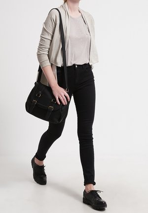 Femme marchant portant un cardigan beige, une chemise rose clair, un jean skinny noir, des chaussures brogues noires, et un sac à bandoulière noir.