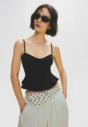 Frau mit kurzen dunklen Haaren trägt schwarze Sonnenbrille, ein schwarzes Spaghettiträgertop, weiße Hose und eine weiße Bauchtasche mit Punkten, Hände in den Taschen.
