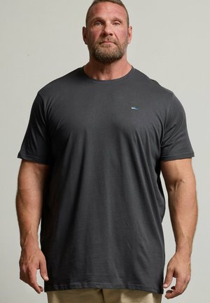 Muskularny mężczyzna z brodą stoi przodem, ma na sobie zwykły ciemnoszary T-shirt i jasne spodnie, na neutralnym tle.