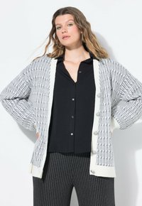 Cardigan grigio e bianco con motivo, caratterizzato da una texture lavorata a maglia, con chiusura a bottoni e maniche larghe, abbinato a una maglietta nera e pantaloni a righe.