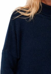 Marineblauer Strickpullover mit hohem Kragen, langen Ärmeln und strukturiertem Finish. Der Stoff wirkt weich und etwas locker sitzend.