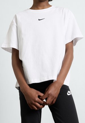 Osoba ubrana w białą koszulkę Nike i czarne spodnie Nike, ręce splecione z przodu, na jednolitym jasnym tle.