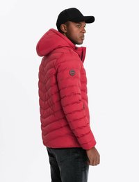Doudoune rouge avec capuche, présentant un design matelassé, un devant zippé et des poches latérales. Accents noirs sur la manche et casquette noire.
