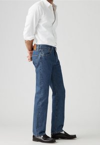 Vaqueros clásicos azules con corte recto, hechos de mezclilla. Presentan una parche de cuero en la cintura trasera y costuras en contraste. Combinados con zapatos negros.