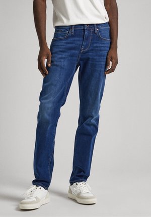 Man draagt blauwe slim-fit jeans, witte sneakers en een wit t-shirt, staande tegen een effen lichte achtergrond.