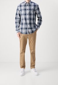 Mężczyzna w granatowo-białej koszuli w kratę, beżowych spodniach i białych trampkach stoi na tle jednolitego białego tła.