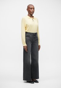 Rumena polo majica z dolgimi rokavi in tremi gumbi, v kombinaciji z visokim pasom in širokimi temno sivimi kavbojkami. Manekenka nosi črne nizke čevlje.