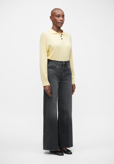 Polo shirt jaune à manches longues avec trois boutons, associé à un jean gris foncé taille haute et à jambes larges. Le modèle porte des chaussures noires à enfiler.