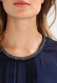 Granatowa bluzka z plisowanym wzorem i koralikowym dekoltem. Koraliki są srebrne, co nadaje szczegóły w okolicy kołnierza.