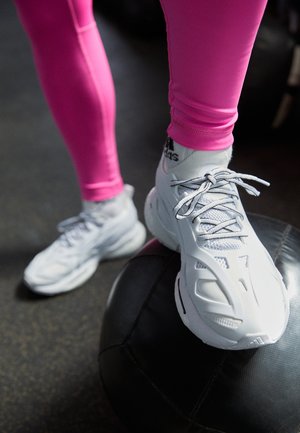 Weiße Sportschuhe aus Mesh und synthetischen Überzügen, mit Schnürsenkeln, geschwungenem Design, die auf einem schwarzen Medizinball ruhen, vor einem dunklen Hintergrund.