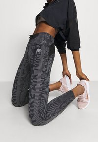 Nike aktiva leggings i svart med grå randiga mönster; hög midja, slätt tyg och logotypdetalj. Matchas med ljusrosa sneakers.