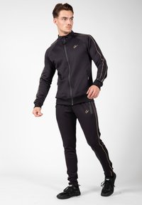 Zwarte sportieve trainingsset met een jack met rits en taps tolopende broek, voorzien van gouden strepen en ritszakken.