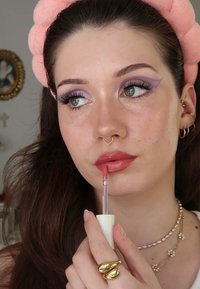 Lucidalabbra con applicatore trasparente, caratterizzato da una tonalità di rosa soft. Gli accessori includono un anello d'oro e collane sovrapposte, su uno sfondo neutro.