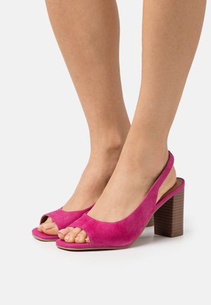 Stopy w jasnoróżowych zamszowych sandałach na obcasie z odkrytymi palcami, z drewnianymi blokami na wysokim obcasie, na białym tle.