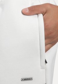 Bílé fleecové tepláky s boční kapsou, s kovovým štítkem s nápisem "SMILODOX." Tkanina má měkkou texturu a čistý design.