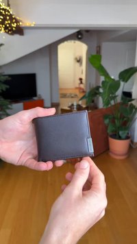 Portefeuille en cuir marron tenu dans une main, présentant une texture lisse, une forme rectangulaire, avec un logo en métal sur un côté.