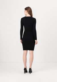 Fekete bordázott ruha hosszú ujjakkal és testhez simuló szabással, térd alá érő szoknyával. Fekete magas sarkú cipővel viselve.