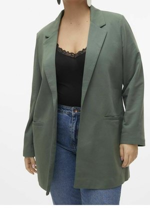 Femme portant un blazer vert olive ouvert, un haut noir bordé de dentelle, un jean bleu et des boucles d'oreilles ovales blanches, sur un fond uni.